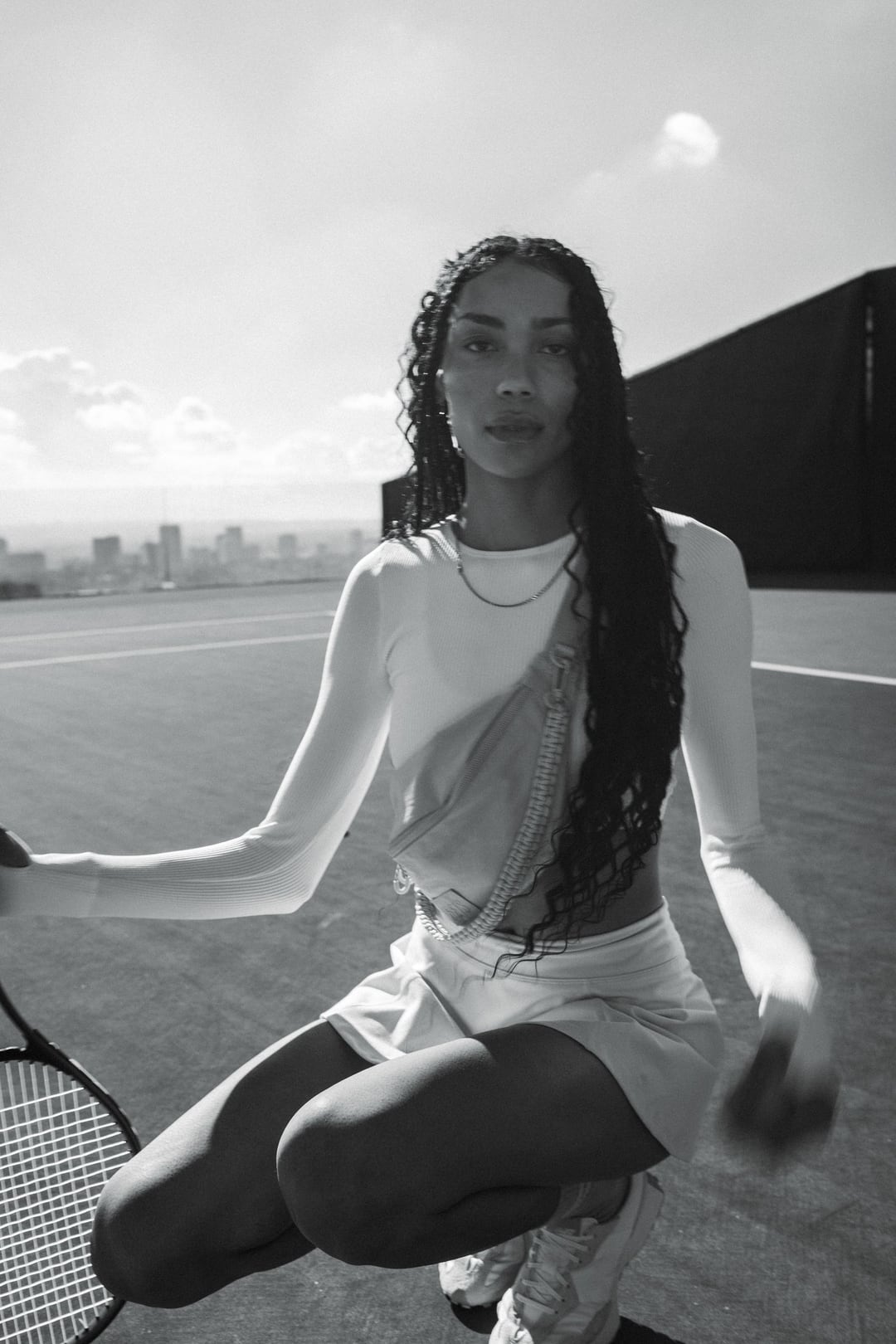 Black and white: a model crouches on a hilltop tennis court, racket in hand, city skyline behind.