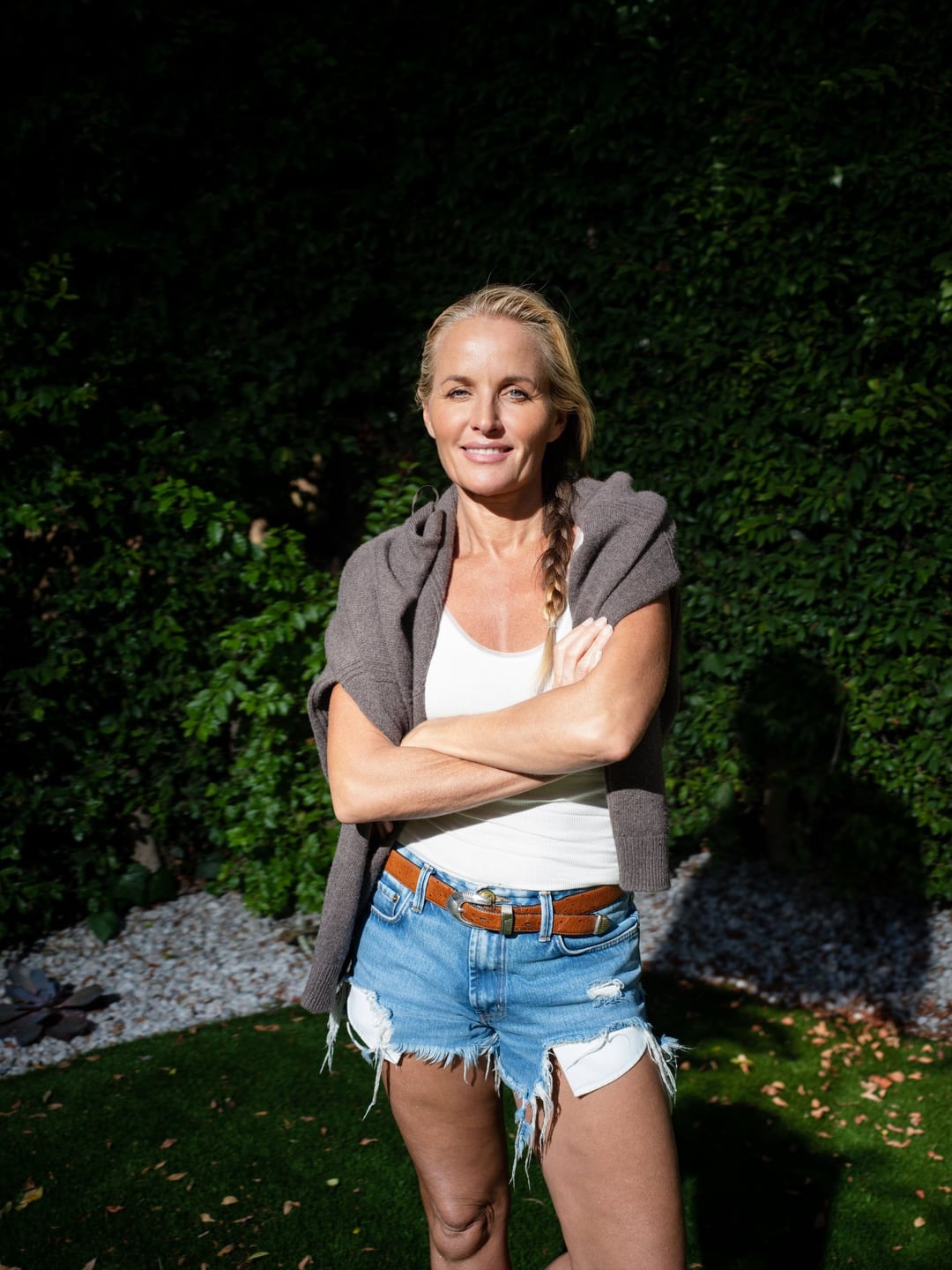 Davinia Taylor waist-up against a dark hedge, arms folded over a white tank, a grey sweater over her shoulders, smiling at the lens.