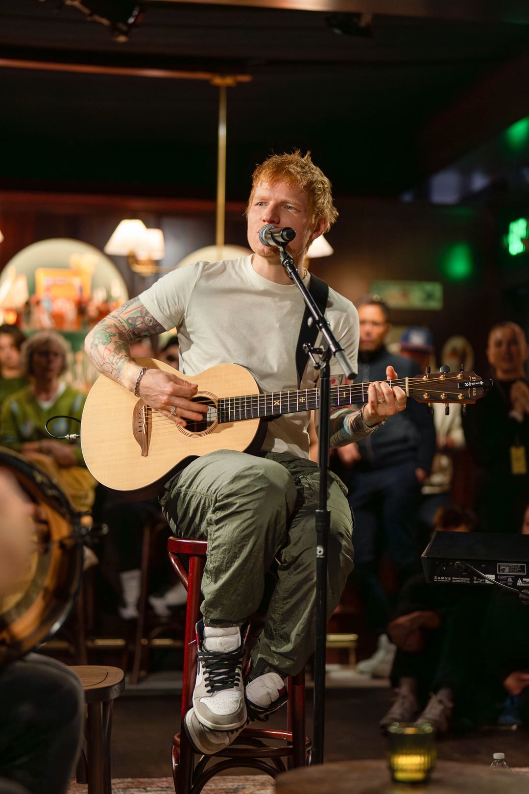 Ed Sheeran singing on a stool with an acoustic guitar in a dark green pub room lit by warm lamps, a crowd behind him.