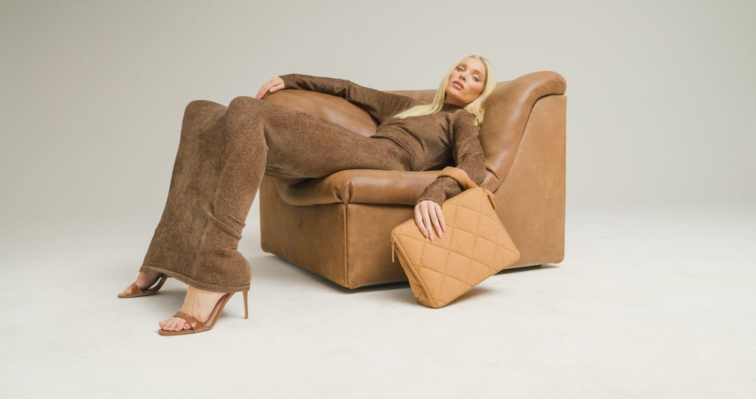 Elsa Hosk lounging sideways across a tan leather armchair in a brown knit set and heels, a quilted tan BÉIS bag in her hand.