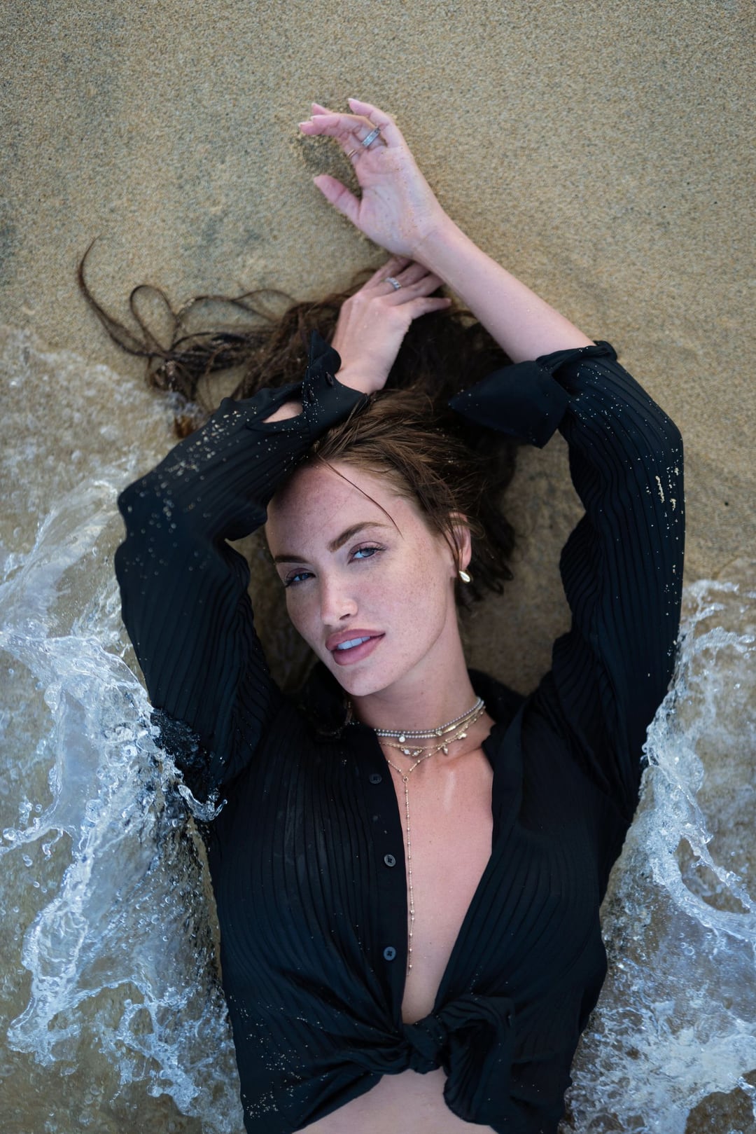 Haley Kalil from above, lying back in shallow surf in an open black shirt, arms over her head.