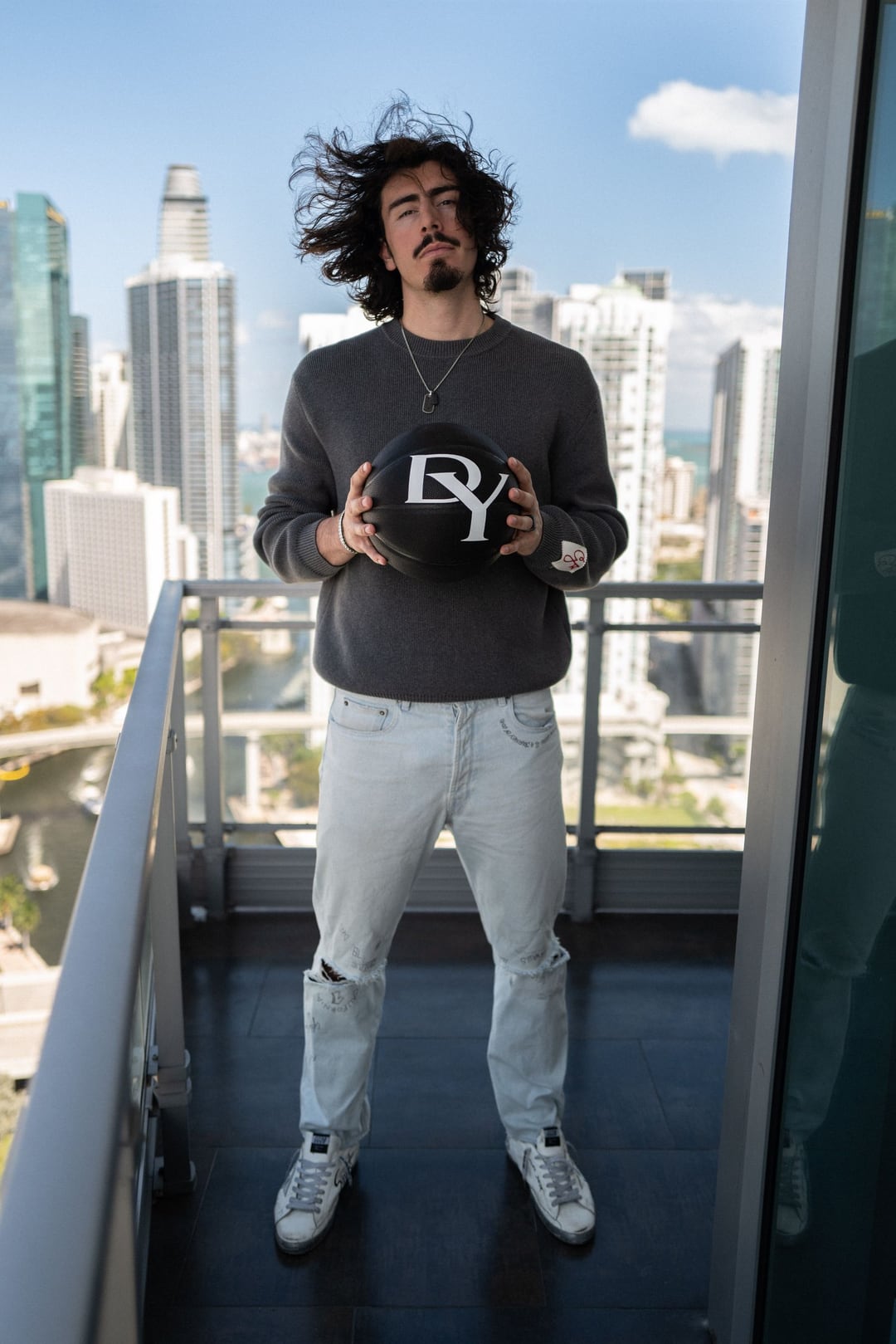 Jaime Jaquez Jr. on a high balcony holding a David Yurman basketball, hair blown up by the wind, city towers behind.