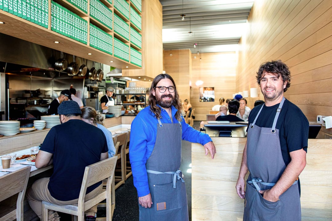 Chefs Jon Shook and Vinny Dotolo in aprons in their restaurant, one in a cobalt shirt, the dining room behind them.