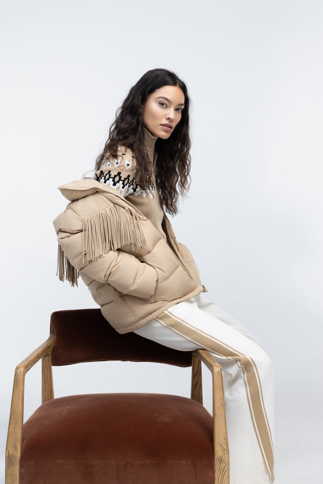 Model seated backwards on a velvet chair in a fringed shearling jacket and cream trousers.
