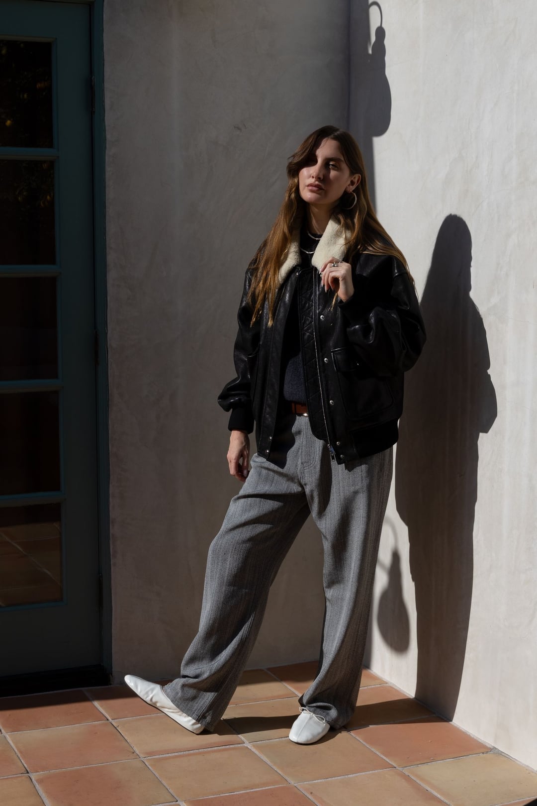 Allison Bornstein in a black leather jacket and grey trousers leaning against a white stucco wall, hard sun casting her shadow.
