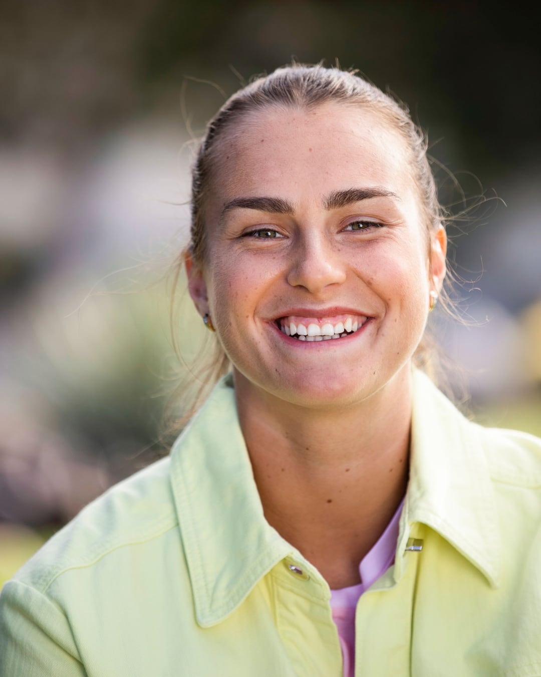Aryna Sabalenka close-up, laughing into the lens in a pale yellow jacket, her hair pulled back.