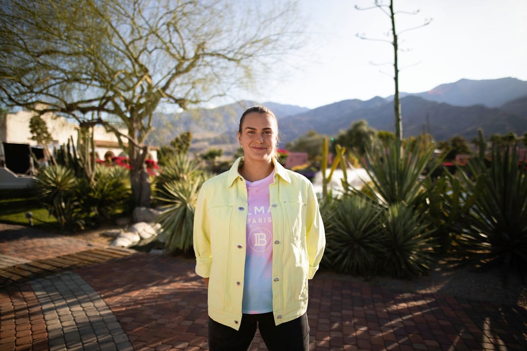 Aryna Sabalenka full length in a pale yellow jacket and black trousers, standing in a desert garden of agaves with mountains behind.